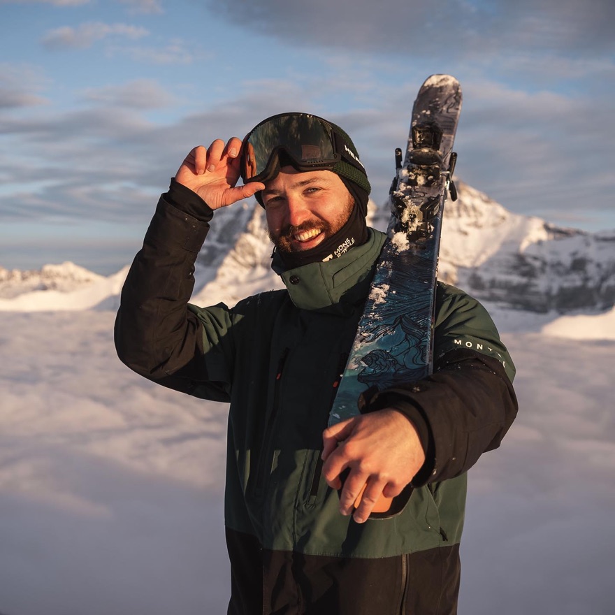 Alex Stuart holding his skis on a mountain top above the clouds